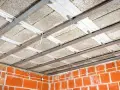 Konstrukcja sufitu podwieszanego z płytami izolacyjnymi i profilem metalowym. Widać ceglane ściany i przewody elektryczne. To etap prac nad suchą zabudową.