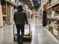 Klient w Castoramie wybiera materiały budowlane, sprawdzając cennik usług. Wózek pełen desek i płyt.