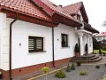 Biała elewacja domu z brązowymi okiennicami i czerwonym dachem. Przed wejściem schody i zadbany ogród.