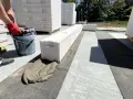 Budowa domu z **suporeksu**. Rękawica z czerwoną rękawicą trzyma wiadro z zaprawą, która jest układana na bloczkach.
