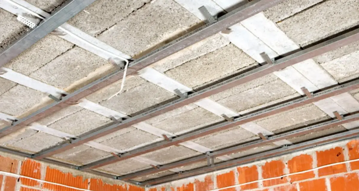Konstrukcja sufitu podwieszanego z płytami izolacyjnymi i profilem metalowym. Widać ceglane ściany i przewody elektryczne. To etap prac nad suchą zabudową.