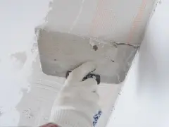 Ręka w rękawicy nakłada zaprawę na styropian przy suficie. Trwa klejenie styropianu do sufitu na piankę.