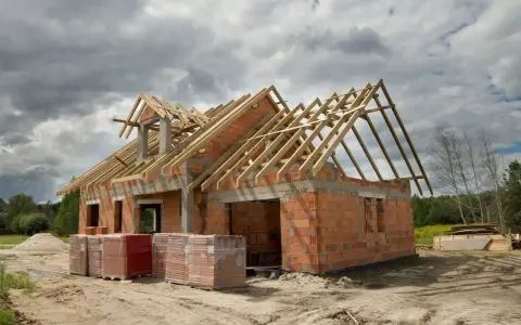 Budowa domu 100m2 w toku. Ściany z cegły, drewniany szkielet dachu, materiały budowlane na placu.