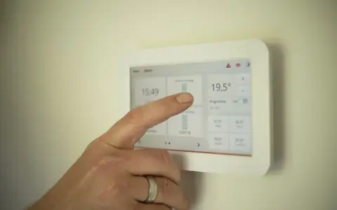 Ręka dotyka ekranu termostatu, sprawdzając, czy piec gazowy powinien chodzić cały czas. Wyświetlana jest temperatura 19,5°C.