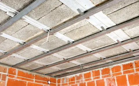 Konstrukcja sufitu podwieszanego z płytami izolacyjnymi i profilem metalowym. Widać ceglane ściany i przewody elektryczne. To etap prac nad suchą zabudową.