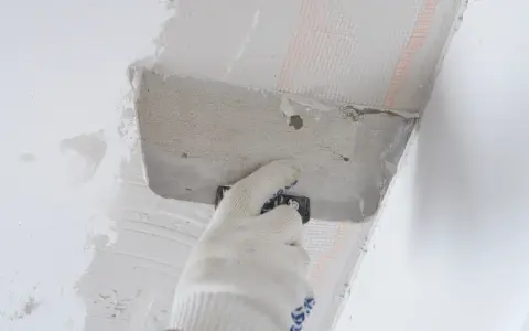 Ręka w rękawicy nakłada zaprawę na styropian przy suficie. Trwa klejenie styropianu do sufitu na piankę.