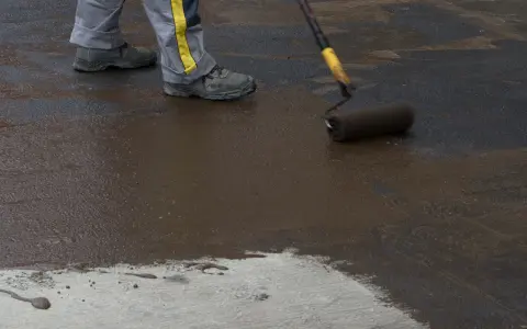 Pracownik wykonuje hydroizolację chudziaka, nakładając masę bitumiczną wałkiem.