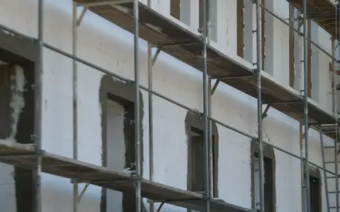 Budowa domu z bloczków styropianowych. Rusztowania otaczają białą ścianę z otworami okiennymi i drzwiami, czekającymi na wykończenie.