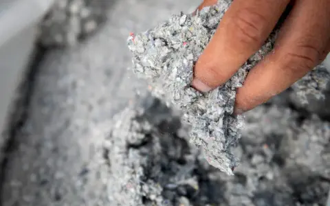 Dłoń trzyma materiał do celuloza ocieplenie. Jest to szara, włóknista masa z kolorowymi drobinkami.
