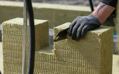 Ręka w rękawicy tnie piłą wełnę skalną 10. Materiał izolacyjny jest przygotowywany do montażu.