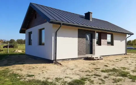 Nowy, nowoczesny dom kanadyjski z ciemnym dachem i białymi ścianami, otoczony zielenią i piaskiem.