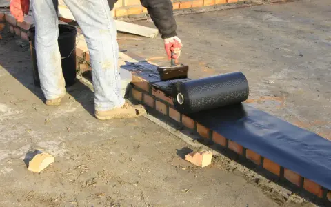 Pracownik kładzie papę na murze, szukając alternatywy dla dysperbitu. Czarna rolka materiału hydroizolacyjnego jest rozwijana na mokrej powierzchni.
