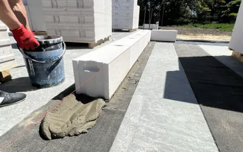 Budowa domu z **suporeksu**. Rękawica z czerwoną rękawicą trzyma wiadro z zaprawą, która jest układana na bloczkach.