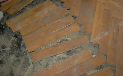 Drewniane deski parkietu, niektóre luźne, czekają na ułożenie. Widać kurz i resztki starej podłogi.