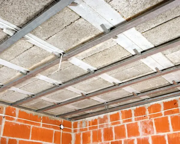 Konstrukcja sufitu podwieszanego z płytami izolacyjnymi i profilem metalowym. Widać ceglane ściany i przewody elektryczne. To etap prac nad suchą zabudową.