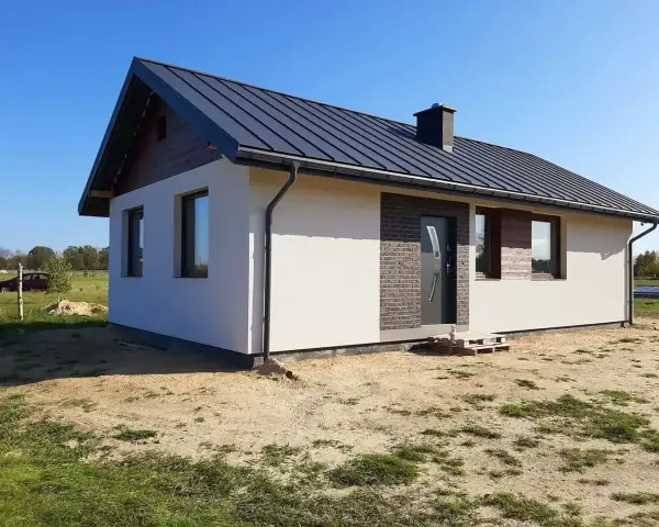 Nowy, nowoczesny dom kanadyjski z ciemnym dachem i białymi ścianami, otoczony zielenią i piaskiem.