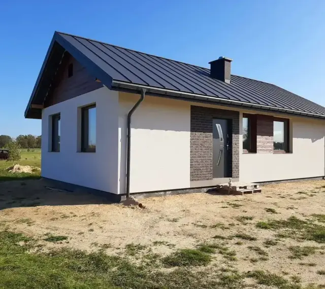 Nowy, nowoczesny dom kanadyjski z ciemnym dachem i białymi ścianami, otoczony zielenią i piaskiem.