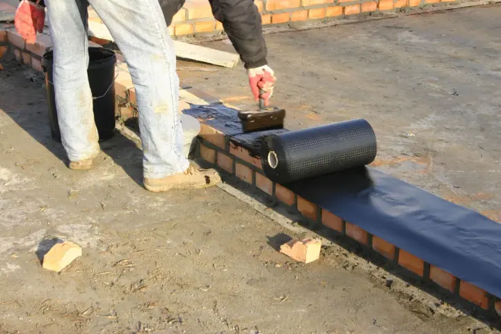 Pracownik kładzie papę na murze, szukając alternatywy dla dysperbitu. Czarna rolka materiału hydroizolacyjnego jest rozwijana na mokrej powierzchni.