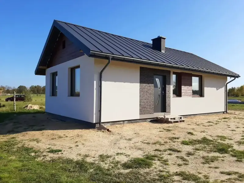 Nowy, nowoczesny dom kanadyjski z ciemnym dachem i białymi ścianami, otoczony zielenią i piaskiem.